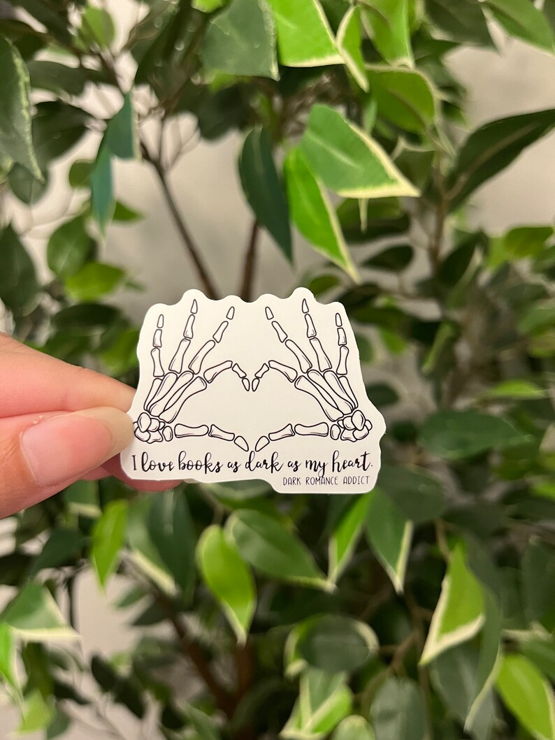 Op de afbeelding: Witte sticker met een zwart skelet hand hart ontwerp. De tekst luidt "I love books as dark as my heart" met "Dark Romance Addict" eronder. De sticker wordt getoond tegen een groene achtergrond.