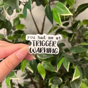 Op de afbeelding: Een witte sticker met de tekst "you had me at TRIGGER WARNING" in een zwarte, vette letter. De sticker wordt voor een achtergrond van groene bladeren gehouden.
