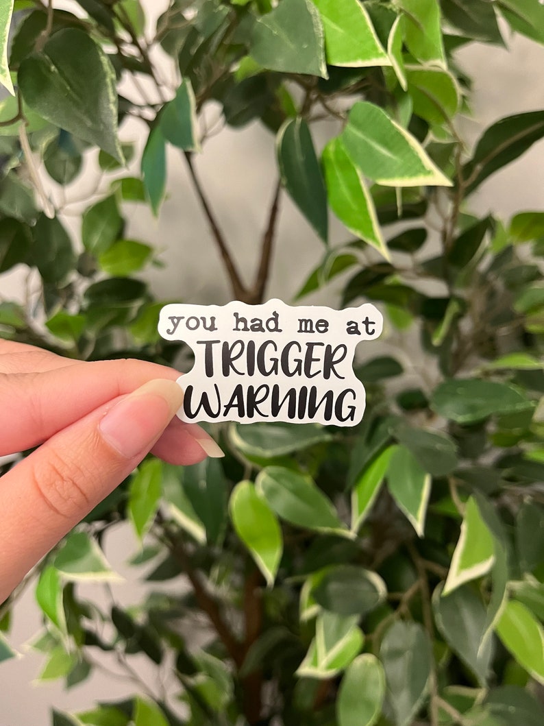 Op de afbeelding: Een witte sticker met de tekst "you had me at TRIGGER WARNING" in een zwarte, vette letter. De sticker wordt voor een achtergrond van groene bladeren gehouden.