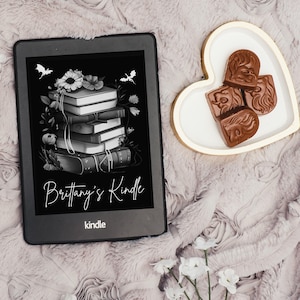 Puede incluir: Una ilustración en blanco y negro de una pila de libros con flores y murciélagos sobre un fondo negro. La imagen se muestra en un lector electrónico Kindle. El texto "Brittany's Kindle" se muestra debajo de la imagen. El lector electrónico Kindle está sobre una superficie rosa peluda con un plato en forma de corazón que contiene cuatro caramelos de chocolate.