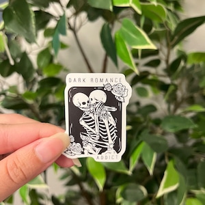 Op de afbeelding: Een zwart-witte sticker met twee omhelzende skeletten, omringd door rozen. De sticker heeft de tekst "DARK ROMANCE" en "ADDICT". De sticker wordt vastgehouden voor een achtergrond van groene bladeren.