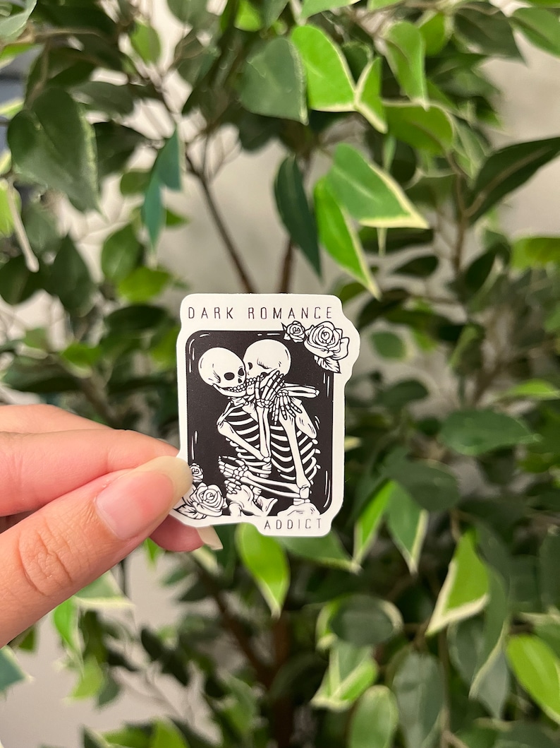 Op de afbeelding: Een zwart-witte sticker met twee omhelzende skeletten, omringd door rozen. De sticker heeft de tekst "DARK ROMANCE" en "ADDICT". De sticker wordt vastgehouden voor een achtergrond van groene bladeren.