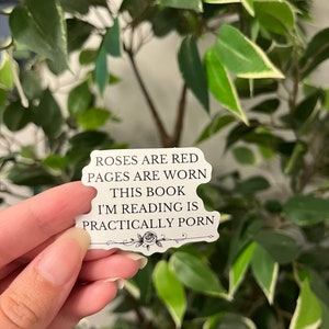 Op de afbeelding: Een witte sticker met zwarte tekst die luidt: "Roses are red, pages are worn, this book I'm reading is practically porn." De sticker heeft een bloemenontwerp aan de onderkant en wordt voor een groene plant met bladeren gehouden.