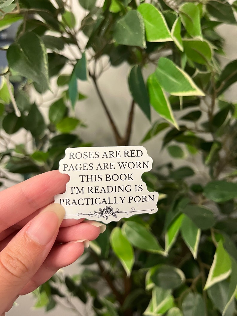 Op de afbeelding: Een witte sticker met zwarte tekst die luidt: "Roses are red, pages are worn, this book I'm reading is practically porn." De sticker heeft een bloemenontwerp aan de onderkant en wordt voor een groene plant met bladeren gehouden.