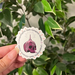 Op de afbeelding: Een witte sticker met een geschulpte rand met een paarse schedel met smeltend was en een brandende kaars. De tekst "Unapologetic dark romance reader" is in roze rond de rand gedrukt. De sticker wordt in een hand gehouden voor een groene, bladerrijke achtergrond.