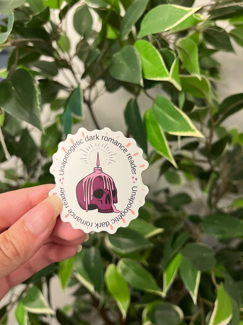 Op de afbeelding: Een witte sticker met een geschulpte rand met een paarse schedel met smeltend was en een brandende kaars. De tekst "Unapologetic dark romance reader" is in roze rond de rand gedrukt. De sticker wordt in een hand gehouden voor een groene, bladerrijke achtergrond.