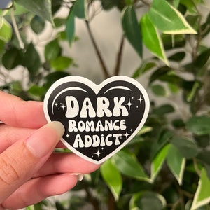 Op de afbeelding: Een zwarte hartvormige sticker met de woorden "DARK ROMANCE ADDICT" in witte, retro letters. De sticker heeft een witte rand en steraccenten. De achtergrond is een vage weergave van groene bladeren.