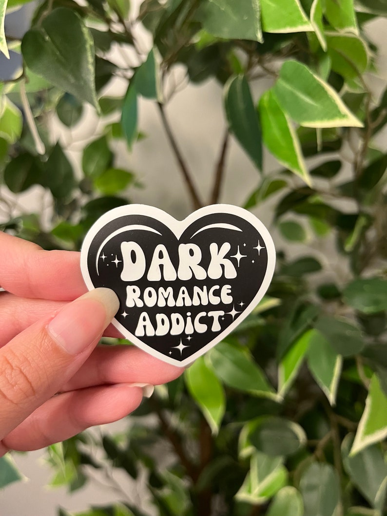 Op de afbeelding: Een zwarte hartvormige sticker met de woorden "DARK ROMANCE ADDICT" in witte, retro letters. De sticker heeft een witte rand en steraccenten. De achtergrond is een vage weergave van groene bladeren.