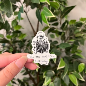 Op de afbeelding: Een zwart-witte sticker met een skelet dat een boek leest, omringd door bladeren. De sticker bevat de tekst "I like 'em real thick and juicy". De sticker wordt gehouden tegen een achtergrond van groene bladeren.