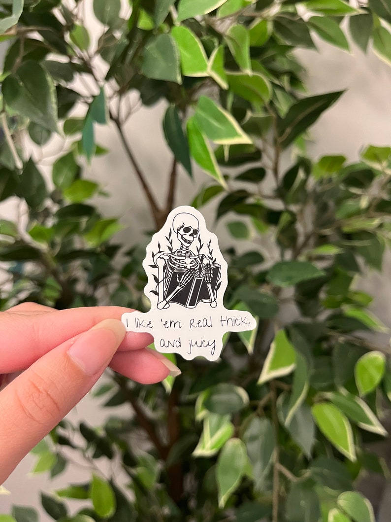 Op de afbeelding: Een zwart-witte sticker met een skelet dat een boek leest, omringd door bladeren. De sticker bevat de tekst "I like 'em real thick and juicy". De sticker wordt gehouden tegen een achtergrond van groene bladeren.