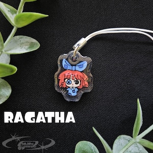 May include: A clear acrylic charm featuring a cartoon doll with red hair, a blue bow, and a blue dress. The charm is attached to a white cord. The word "RAGATHA" is printed in white below the charm.