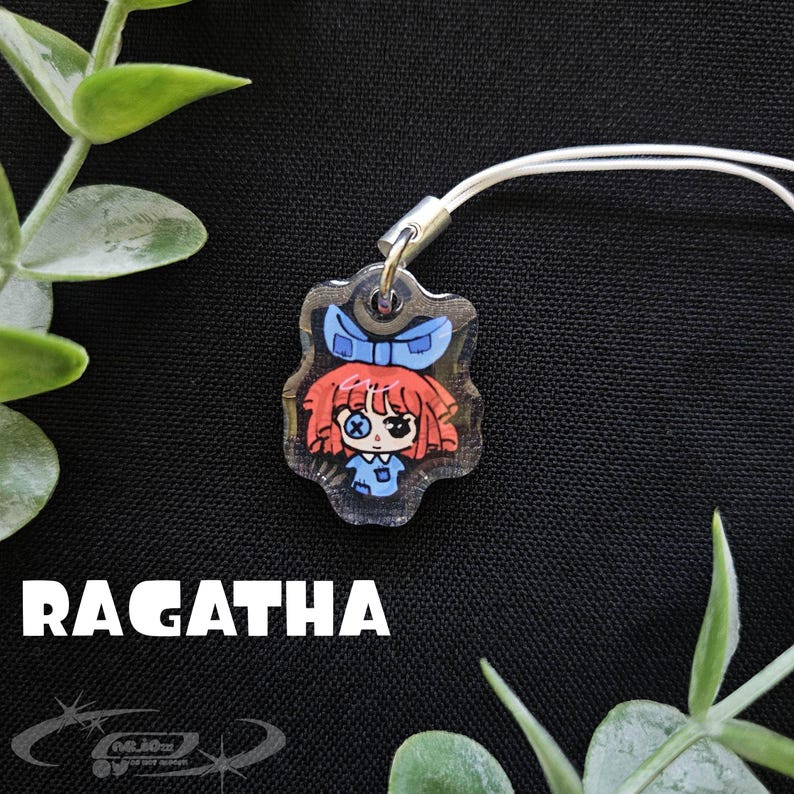 May include: A clear acrylic charm featuring a cartoon doll with red hair, a blue bow, and a blue dress. The charm is attached to a white cord. The word "RAGATHA" is printed in white below the charm.