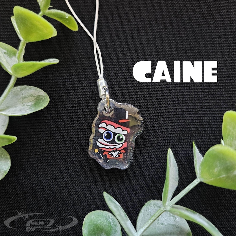 May include: A clear acrylic charm with a cartoon character design. The character has a red and black outfit, a top hat, and large blue eyes. The word "CAINE" is printed in white block letters. The charm is attached to a white cord.