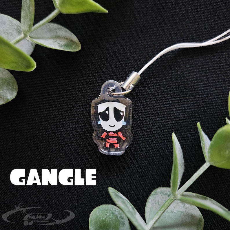 May include: A clear acrylic charm with a cartoon character design. The character has a white face with black eyes and a sad expression, with red accents on the body. The word "GANGLE" is printed in white. A white cord is attached to the charm.