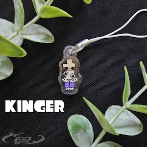 May include: A clear acrylic charm featuring a cartoon character with a cross on its head, blue eyes, and purple pants. The charm is attached to a white cord. The word "KINGER" is printed in white block letters.