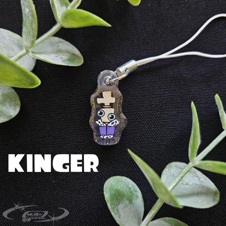 May include: A clear acrylic charm featuring a cartoon character with a cross on its head, blue eyes, and purple pants. The charm is attached to a white cord. The word "KINGER" is printed in white block letters.