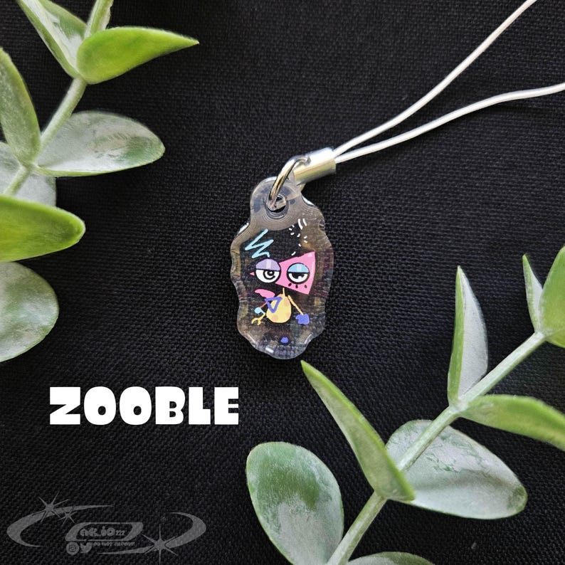 May include: A clear acrylic charm with a cartoon character design. The character has pink, blue, and yellow accents, with the word "ZOOBLE" in white block letters below. The charm is attached to a white cord.