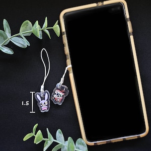 May include: Two clear acrylic phone charms with cartoon designs, one a bunny and the other a mummy. The bunny charm is 1.5 inches tall. Both charms have white cords attached. A smartphone in a gold case is in the background.