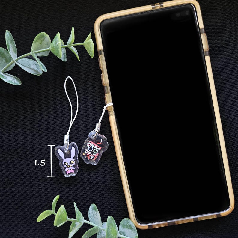 May include: Two clear acrylic phone charms with cartoon designs, one a bunny and the other a mummy. The bunny charm is 1.5 inches tall. Both charms have white cords attached. A smartphone in a gold case is in the background.