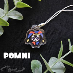 May include: A clear acrylic charm featuring a cartoon jester character with a red, blue, and yellow hat and outfit. The charm is attached to a white cord. The word "POMNI" is printed in white block letters below the charm.