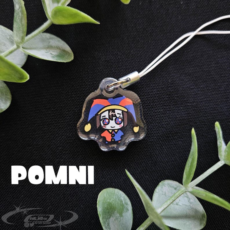 May include: A clear acrylic charm featuring a cartoon jester character with a red, blue, and yellow hat and outfit. The charm is attached to a white cord. The word "POMNI" is printed in white block letters below the charm.