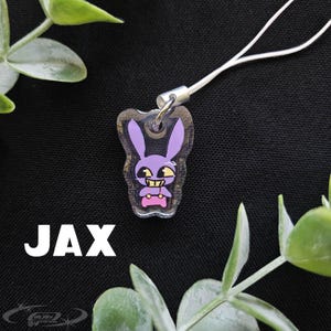 May include: A clear acrylic charm featuring a purple cartoon bunny with yellow eyes and a pink controller. The charm is attached to a white cord and has the name "JAX" in white block letters. The background is black with green foliage.