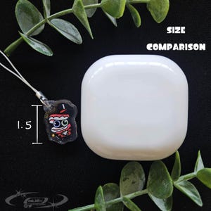 May include: A white, rounded square object next to a small, clear charm with a cartoon character design. The charm is 1.5 inches in height. The text "SIZE COMPARISON" is above the white object.