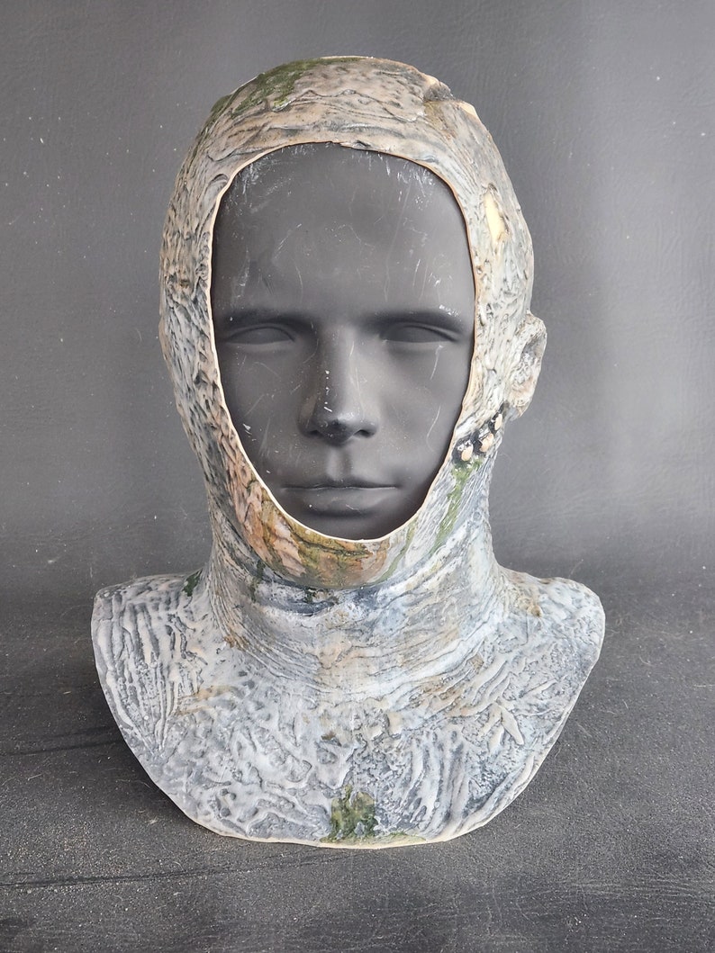 K&ouml;nnte beinhalten: Eine Vollkopfmaske mit Halsst&uuml;ck, pr&auml;sentiert auf einem Schaufensterkopf. Die Maske hat ein Tarnmuster in Grau-, Braun- und Gr&uuml;nt&ouml;nen, das ein verwittertes Aussehen erzeugt. Die Maske hat eine &Ouml;ffnung f&uuml;r das Gesicht.