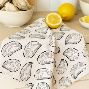 以下が含まれることがあります： 黒いカキの殻模様の白いキッチンタオル、45.7 x 53.3 cm。画像にはレモン、カキのボウル、その他の台所用品も写っています。
