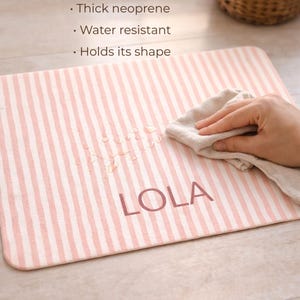 Puede incluir: Una alfombra rectangular a rayas rosas y blancas con el nombre "LOLA" impreso en rosa a juego. La alfombra se est&aacute; limpiando con un pa&ntilde;o beige. El texto en la alfombra dice que es de neopreno grueso, resistente al agua y mantiene su forma.