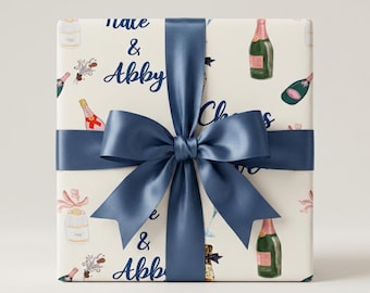 Pode incluir: Uma caixa de presente quadrada embrulhada em papel creme com um padrão repetitivo de garrafas e taças de champanhe, com o texto "Nate & Abby" impresso em letra cursiva azul-escura, atada com um laço de fita de cetim azul-escuro no centro.