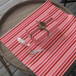 May include: A rectangular gift box wrapped in paper with vertical red, light pink, and dark pink stripes, tied with a brown ribbon bow. A roll of clear adhesive tape and a pair of silver metal scissors are placed on the striped wrapping paper.