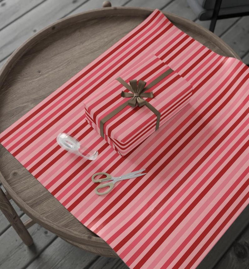 May include: A rectangular gift box wrapped in paper with vertical red, light pink, and dark pink stripes, tied with a brown ribbon bow. A roll of clear adhesive tape and a pair of silver metal scissors are placed on the striped wrapping paper.