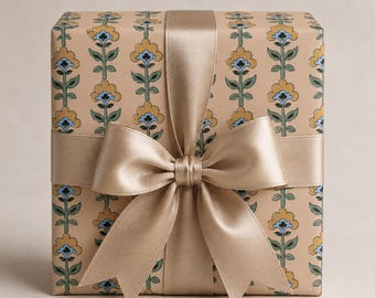 Puede incluir: Una caja de regalo rectangular envuelta en papel de color canela claro con un patrón vertical repetitivo de flores amarillas estilizadas con centros azules y tallos verdes. Una cinta de satén ancha y lisa color champán forma un gran lazo frontal.