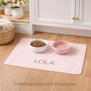 Puede incluir: Una alfombrilla para la alimentaci&oacute;n de mascotas a rayas rosas y blancas con el nombre "LOLA" impreso. La alfombrilla contiene un cuenco blanco con comida para mascotas y un cuenco rosa con agua. El texto "A feeding space you'll actually love" est&aacute; en la parte inferior.