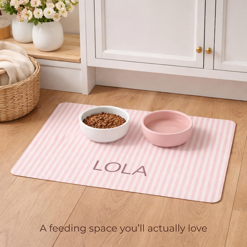 Puede incluir: Una alfombrilla para la alimentaci&oacute;n de mascotas a rayas rosas y blancas con el nombre "LOLA" impreso. La alfombrilla contiene un cuenco blanco con comida para mascotas y un cuenco rosa con agua. El texto "A feeding space you'll actually love" est&aacute; en la parte inferior.