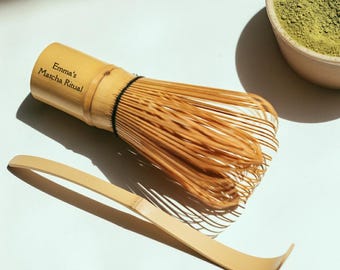 May include: A bamboo matcha whisk, scoop, and a bowl of green matcha powder are displayed on a white surface. The whisk has the text "Emma's Matcha Ritual" printed on the handle. The whisk and scoop are light brown.