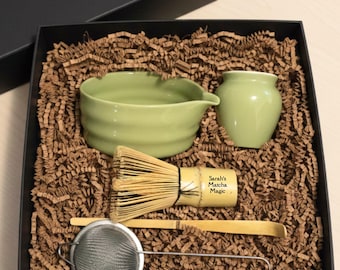 May include: A matcha tea set in a black gift box. The set includes a green ceramic bowl, a small green jar, a bamboo whisk, a bamboo scoop, and a stainless steel sieve. The whisk handle has the text "Sarah's Matcha Magic".