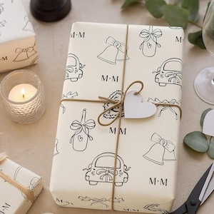 May include: A cream-coloured gift wrapped in paper printed with black line drawings of bells, cars, and bows. The package is tied with twine and a white heart-shaped tag. The initials "M+M" are printed on the wrapping paper.