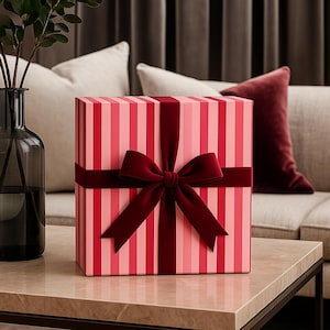May include: A square gift box featuring a vertical striped pattern in alternating shades of light pink and dark pink. The box is wrapped with a wide, dark red velvet ribbon tied into a prominent bow centred on the front face of the package.