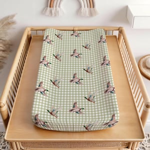 Könnte beinhalten: Eine Baby-Wickelauflage mit einem grün-weißen Vichy-Muster. Die Auflage ist mit Aquarellillustrationen von fliegenden Enten verziert. Sie liegt auf einem hellbraunen Wickeltisch.