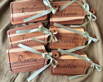 Puede incluir: Una colección de tablas de cortar de madera personalizadas. Cada tabla es rectangular con un asa y presenta un nombre grabado en cursiva. Cintas verde claro están atadas alrededor de las asas. Las tablas tienen una veta de madera natural.