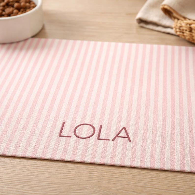Puede incluir: Alfombrilla para comida de mascotas a rayas rosas y blancas con el nombre "LOLA" bordado en hilo rosa a juego. La alfombrilla es rectangular y est&aacute; hecha de tela texturizada. Un cuenco blanco con comida para mascotas es visible en el fondo.
