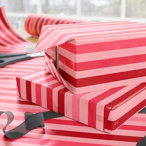 May include: Two rectangular boxes wrapped in glossy paper featuring horizontal stripes in alternating shades of red, dark pink, and light pink. A roll of matching striped wrapping paper, a pair of black scissors, and a black ribbon are visible.