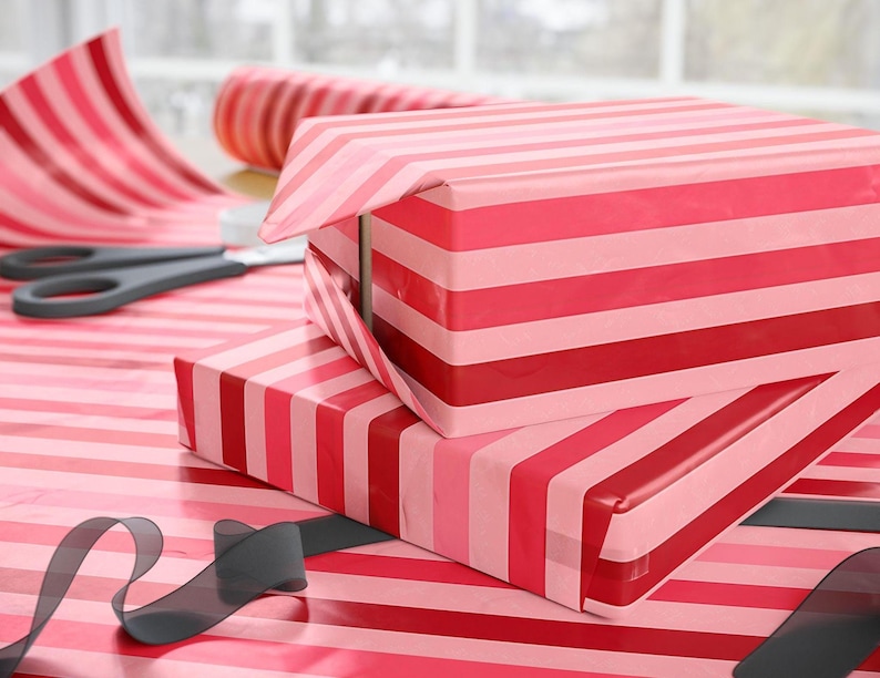 May include: Two rectangular boxes wrapped in glossy paper featuring horizontal stripes in alternating shades of red, dark pink, and light pink. A roll of matching striped wrapping paper, a pair of black scissors, and a black ribbon are visible.