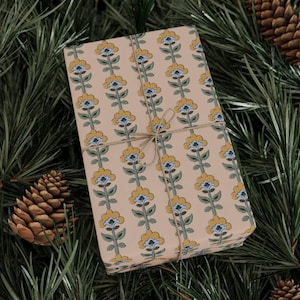 May include: A wrapped gift with floral patterned wrapping paper. The paper features a repeating vertical design of yellow and blue flowers on a tan background. The gift is tied with twine and rests on a bed of green pine needles and pine cones.