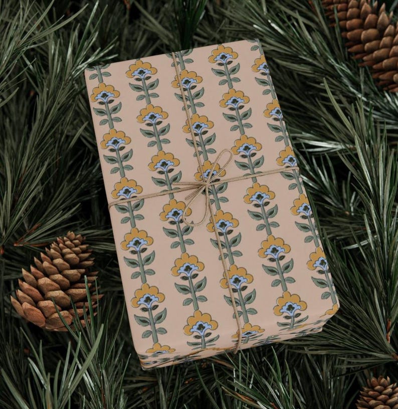 May include: A wrapped gift with floral patterned wrapping paper. The paper features a repeating vertical design of yellow and blue flowers on a tan background. The gift is tied with twine and rests on a bed of green pine needles and pine cones.