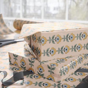 May include: Two gift boxes wrapped in floral patterned paper with yellow, blue, and green accents. A roll of matching paper and scissors are visible in the background. A gray ribbon is draped across the bottom.