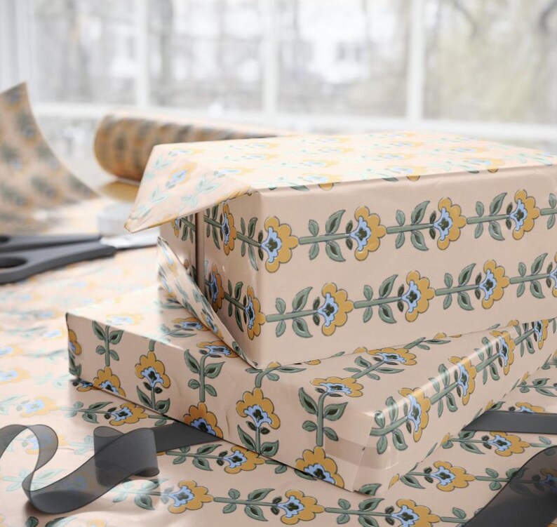 May include: Two gift boxes wrapped in floral patterned paper with yellow, blue, and green accents. A roll of matching paper and scissors are visible in the background. A gray ribbon is draped across the bottom.