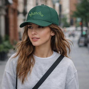 May include: A dark green baseball cap with the word "Matcha" embroidered in a light green script across the front. The cap has a curved brim and a classic six-panel design. The background is blurred, suggesting an outdoor setting.